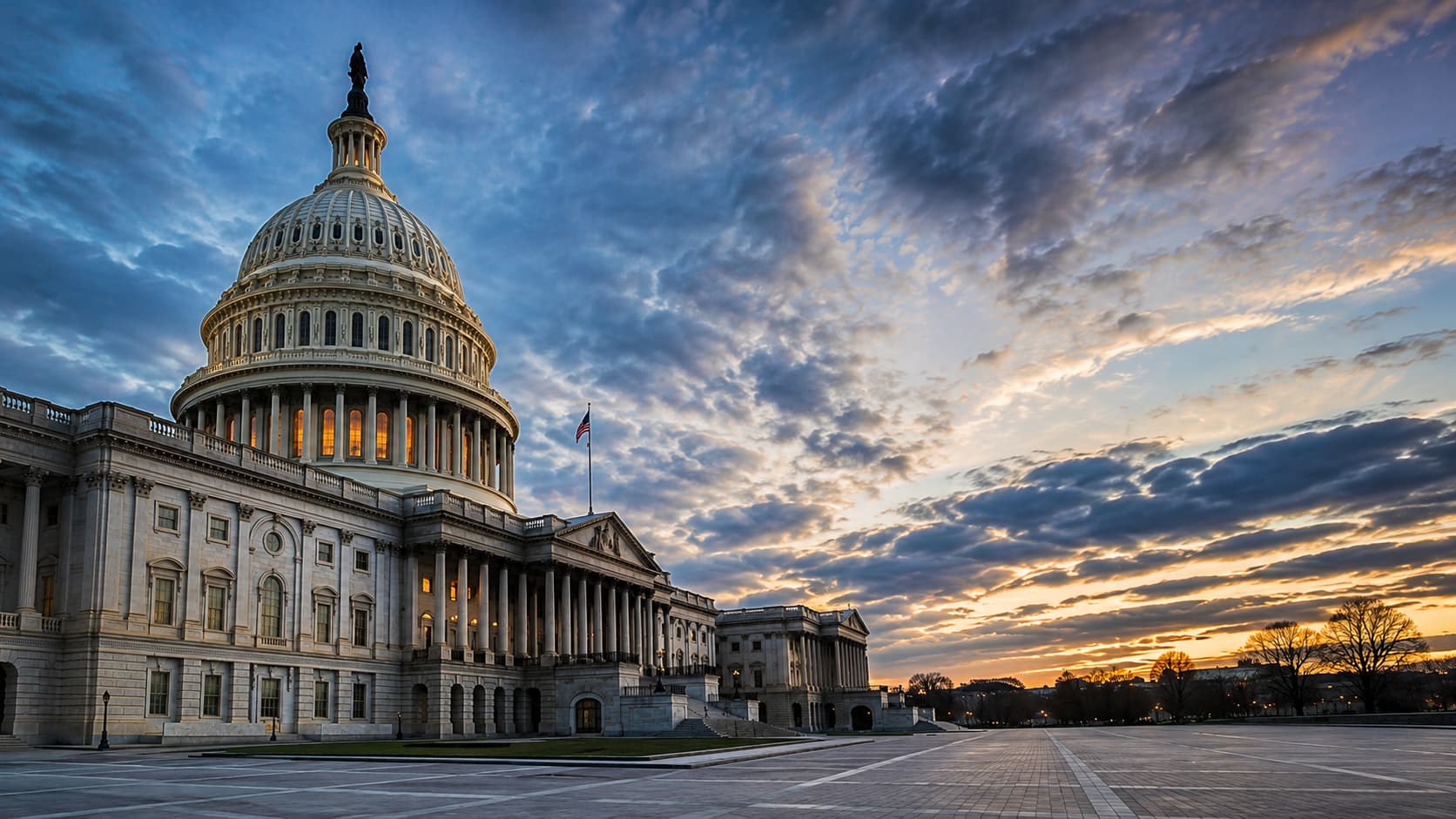 Wide-angle editorial photograph of the U.S. Capitol building at dusk, viewed from a low upward angle with the Capitol positioned left of center. The sky is filled with dramatic blue-gray clouds and warm amber light near the horizon, creating a cinematic, slightly moody atmosphere. The stone architecture is softly illuminated with glowing interior windows, while the expansive empty plaza on the right provides negative space for text. No people are present.
